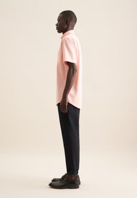 Camicia a maniche corte di color rosa chiaro con taschino sul petto, abbinata a pantaloni tapered scuri e scarpe di pelle nere, vista di profilo.