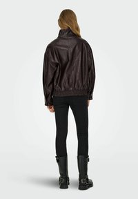 Giubbotto di pelle marrone con colletto alto, orlo elastico e vestibilità oversize. Indossato con jeans skinny neri e stivali neri alti fino al ginocchio.