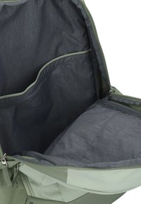 Grünes Stoff-Rucksackinnere, ausgestattet mit einem dunkelgrauen Futter, einer flachen Tasche und einem gepolsterten Fach. Reißverschlussöffnung entlang der Oberseite.