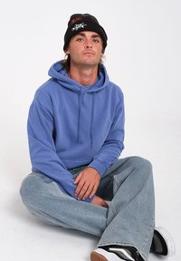 Felpa blu in cotone con coulisse, polsini a coste e una tasca frontale, abbinata a jeans a gamba larga di un blu chiaro e sneaker nere. Berretto nero con grafiche.