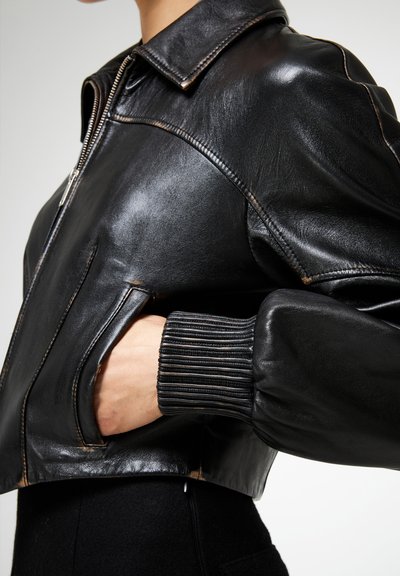 Chaqueta bomber de cuero negra con cuello, cierre de cremallera, puños de canalé y bolsillos laterales. El material tiene una textura lisa y ligeramente brillante.