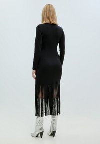 Schwarzes, langärmliges Kleid mit Fransenverzierung am Saum, das eine taillierte Silhouette hat. Kombiniert mit metallic-silbernen, hochhackigen Stiefeln.