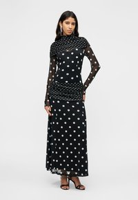 Μακρύ μαύρο φόρεμα με λευκές πουά, διαφανή μακριά μανίκια, σούρες στη μέση και ψηλό λαιμόκομμα. Συνδυάζεται με μαύρα σανδάλια με λουράκια.