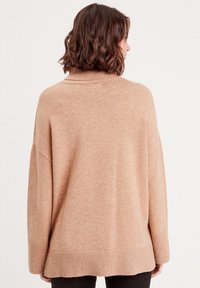 Pull beige oversize avec col roulé, poignets et ourlet côtelés. Texture douce, silhouette décontractée et épaules tombantes. Pas de motifs visibles.