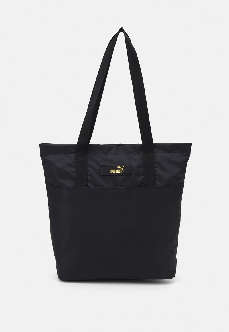 Sac fourre-tout noir en tissu résistant avec deux bandoulières noires. Présente un patch logo doré à l'avant, design minimaliste.
