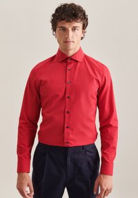 Chemise en coton rouge avec un col classique, des manches longues et des boutons navy, associée à un pantalon bleu foncé. Texture lisse, design ajusté.