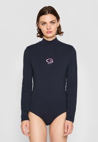 Marineblauwe longsleeve bodysuit van katoenmix. Heeft een hoge hals en een geborduurd "G"-logo in roze en wit op de borst.