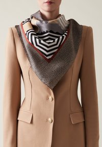 Blazer beige con bottoni in metallo dorato, abbinato a un foulard di seta decorato con design geometrici in colori e texture tenui.