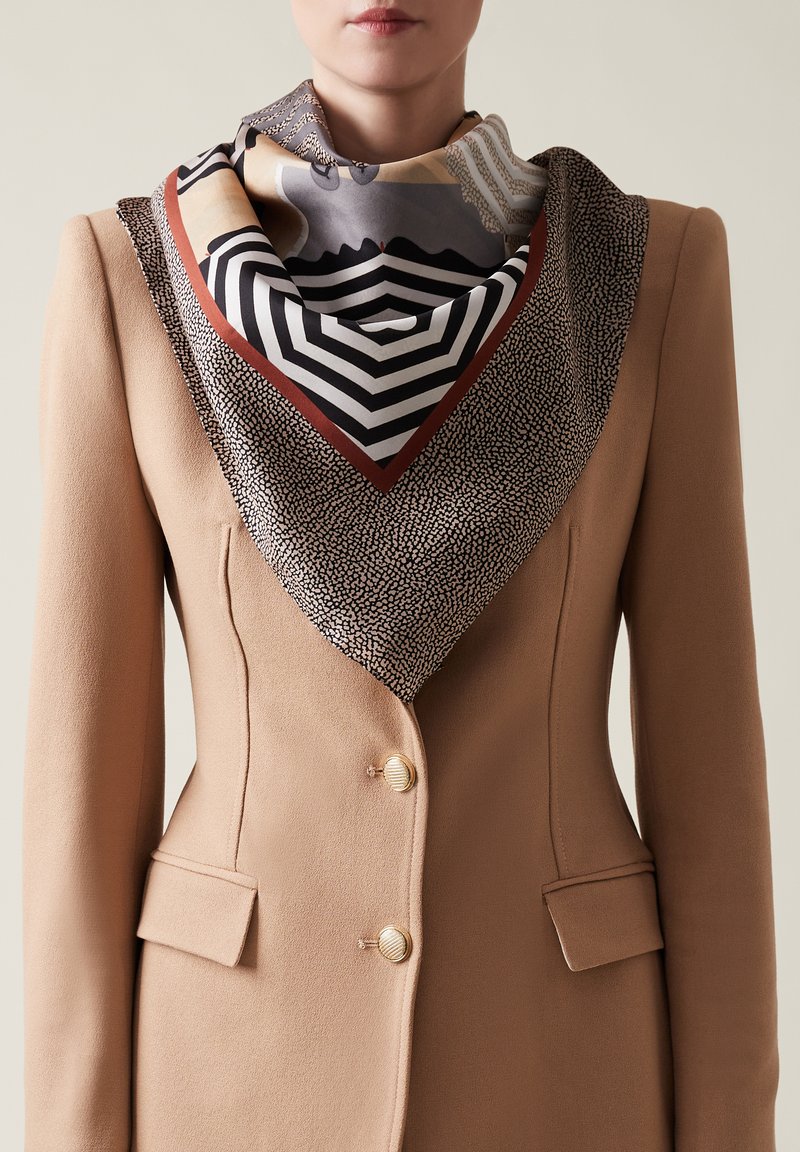 Blazer beige con bottoni in metallo dorato, abbinato a un foulard di seta decorato con design geometrici in colori e texture tenui.