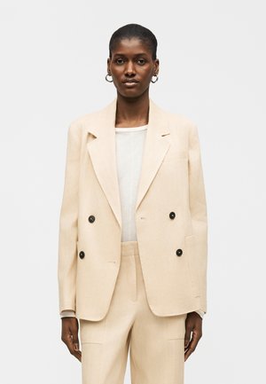 Žena v béžovom blejzri s dvojradovým zapínaním a zodpovedajúcich nohaviciach, na bielom tričku, stojaca pred jednoduchým svetlým pozadím.