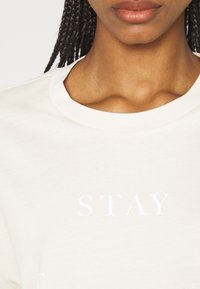 Krémové tričko s kulatým výstřihem, které má výrazný bílý nápis "STAY." Měkký bavlněný materiál s hladkou texturou.