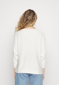 Cremfarbener, strukturierter Pullover mit Dreiviertelärmeln, rundem Ausschnitt und lässiger Passform, kombiniert mit hellblauen Jeans.