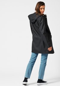 Manteau imperméable noir avec capuche, longueur genoux, boutons-pression et poches latérales. Porté sur un jean bleu clair et des chaussures en toile noires.