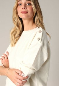 Pull en crème, texturé, avec des épaules tombantes, présentant des boutons décoratifs sur l'épaule. Matière douce, coupe décontractée et poignets côtelés.