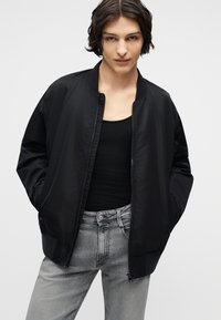 Νεαρός με σκούρα κυματιστά μαλλιά φοράει μαύρο μπουφάν τύπου bomber πάνω από μαύρο τοπ και ανοιχτό γκρι τζιν, με τα χέρια στις τσέπες του μπουφάν.