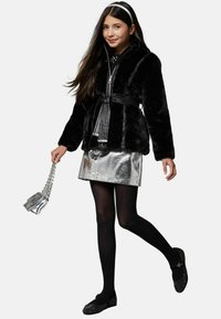 Schwarze Kunstpelzjacke mit einem gebundenen Gürtel, glänzender silberner Minirock, schwarze Strumpfhosen und verzierte schwarze Schuhe, die eine kleine silberne Handtasche hält.