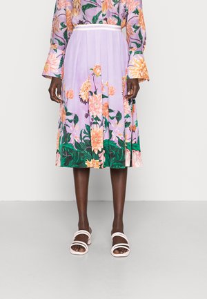 Średniej długości lawendowa spódnica z dużym brzoskwiniowo-zielonym kwiatowym wzorem, zestawiona z pasującą kwiatową bluzką z długim rękawem oraz białymi sandałami na ramiączkach.