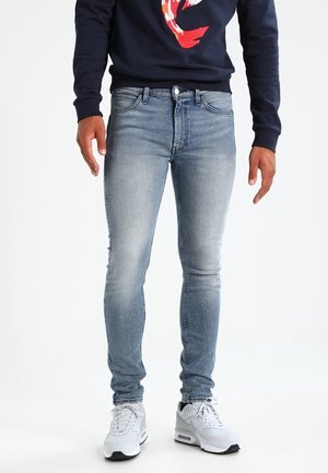 Mand iført lyseblå skinny jeans, hvide sneakers og en marineblå sweatshirt med et rødt og hvidt koi-fisk design.