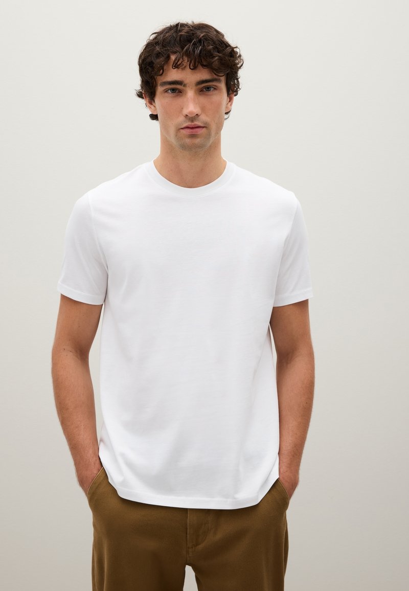 Giovane uomo con capelli ricci, che indossa una maglietta bianca a maniche corte e pantaloni marroni, in piedi con le mani in tasca su uno sfondo chiaro.