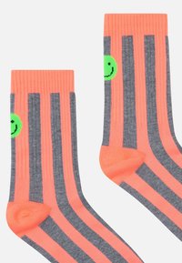 Värikkäät raidalliset sukat, joissa kirkkaan neonpinkkiä ja harmaata kangasta, sivussa pieni vihreä hymiökuvio.