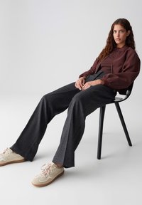 Young woman with wavy hair sits on black chair, wearing brown jacket, dark wide-leg trousers, and beige sneakers against gray background.