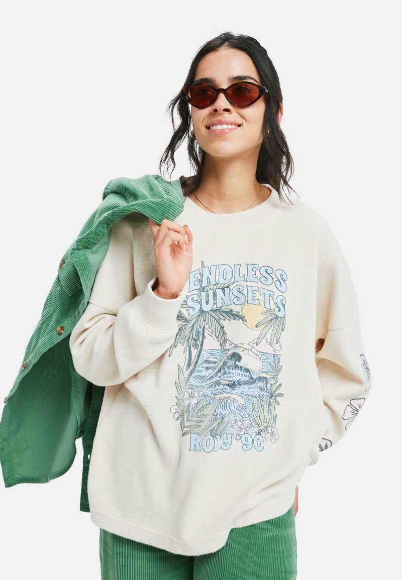 Kerman neulepaita, jossa on "Endless Sunsets" -grafiikka ja palmukuvio, yhdistettynä vihreään corduroytakkia ja -housuja; aurinkolasit.