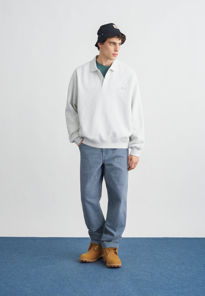 Junger Mann mit schwarzem Anglerhut, hellgrauem Pullover, blauer Hose und hellbraunen Arbeitsstiefeln, der auf einem blauen Teppich vor einer weißen Wand steht.