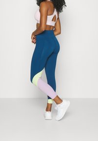 Žena stojící v modrých, levandulových a limetkových legínách s barevnými bloky, s světle růžovým sportovním podprsenkou a bílými teniskami na jednobarevném pozadí.