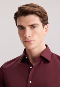 Bordeaux knoopshirt met een puntige kraag, voorzien van witte knopen en een glad, reflecterend oppervlak. Close-up van het bovenlichaam.