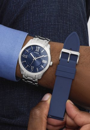 Rozsdamentes acél óra kék számlappal, amely római számokat és ezüst díszítéseket tartalmaz. Az óra kék gumiszíjjal rendelkezik, melyet kézben tartanak.