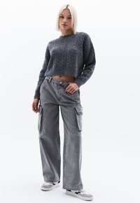 Grauer, kabelgestrickter, verkürzter Pullover mit gerippten Ärmeln, kombiniert mit lockeren, grauen Cargo-Hosen mit seitlichen Taschen und einem dezenten Texturmuster.