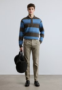 Μακρυμάνικο πόλο με ρίγες σε μπλε και καφέ, συνδυασμένο με μπεζ παντελόνι. Το μοντέλο κρατά μια στρογγυλή μαύρη τσάντα. Λείο ύφασμα, casual εφαρμογή.