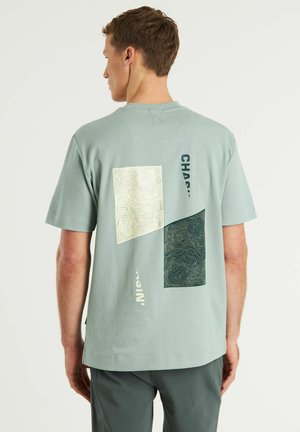 Lichtgroene katoenen t-shirt met abstracte witte en donkergroene geometrische vormen met lijnpatronen en tekst op de achterkant.
