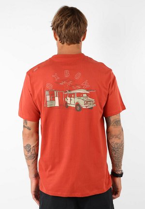 T-shirt en coton rouge avec un graphique d'une camionnette et d'un abri au dos. Manches courtes, col rond, coupe décontractée et tatouages visibles sur les bras.