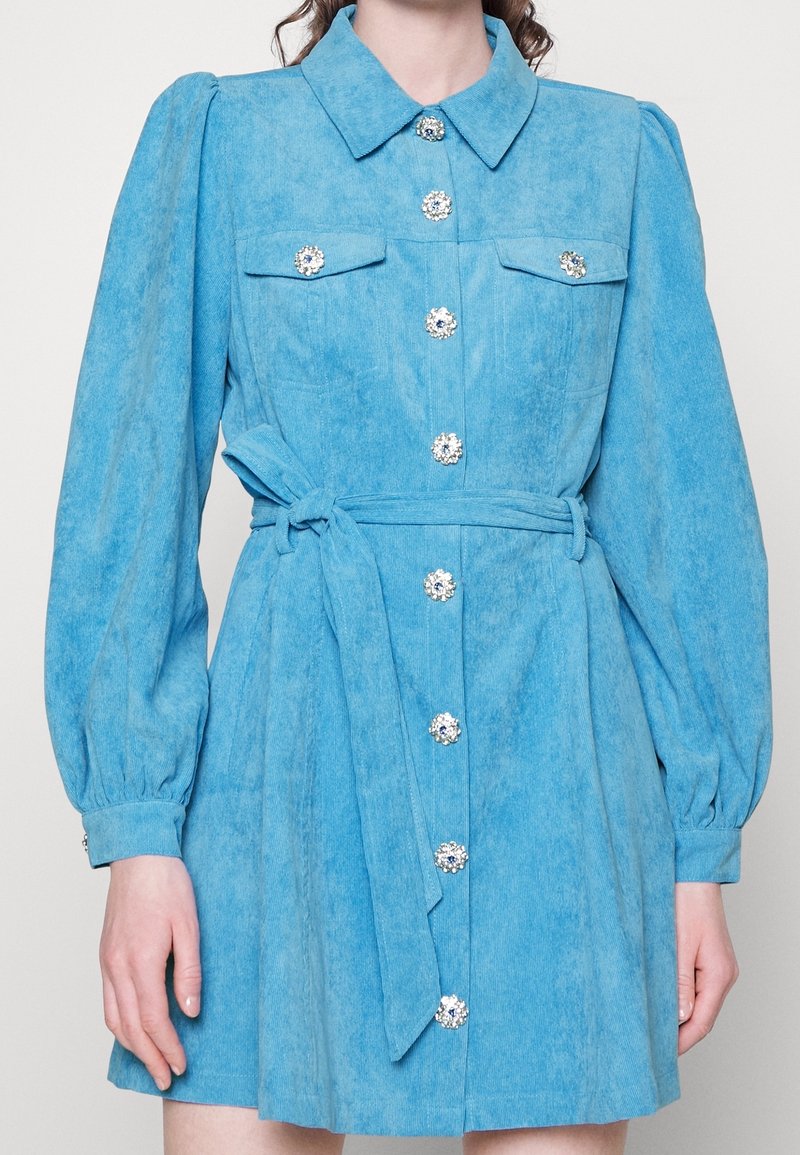 Robe bleue à manches longues avec col, boutons décoratifs en pierres précieuses sur le devant, poches poitrine et ceinture à nouer à la taille.