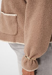 Bruine, textuurvolle fleece jas met witte stiksel op de zak en manchet, gedragen met een gouden armband en lichtgrijze broek.