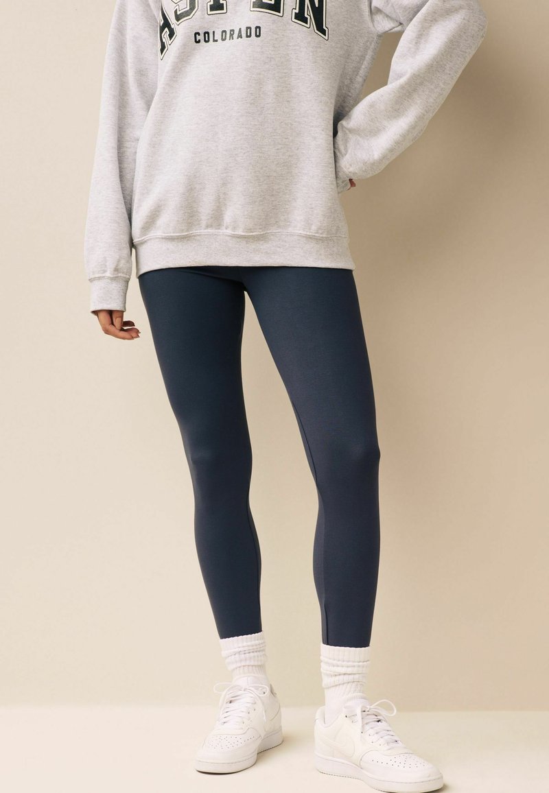 Sudadera gris con el texto "ASPEN COLORADO", combinada con leggings azul oscuro y zapatillas blancas. La sudadera tiene un corte holgado, mientras que los leggings son ajustados.