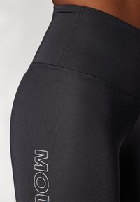 Leggings pretos de cintura alta feitos de um tecido elástico, com costuras planas e um logótipo branco na coxa esquerda. Textura suave.