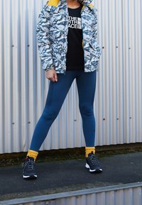 Osoba oblečená v modré maskáčové bundě, černém tričku The North Face, modrých legínách, žlutých ponožkách a černých turistických botách, stojí s nohama rozkročenýma.