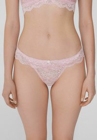 Pink lace thong with floral pattern, scalloped edges, and a small button detail at the front. Soft, delicate texture. Illustrates intricate design.