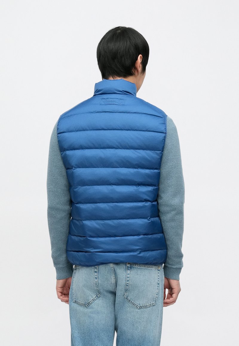 Blue puffer vest with quilted design, high collar, and a smooth finish, worn over a gray long-sleeve shirt and blue jeans.