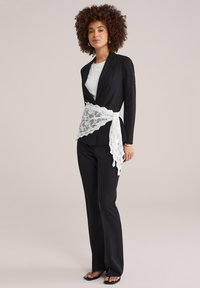 Kobieta o kręconych włosach, ubrana w czarny garnitur, biały top, czarne sandały oraz białą koronkową chustę zawiązaną wokół talii, stojąca na jednolitym tle.