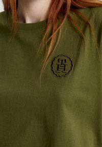 Olivgrön bomulls-T-shirt med en präglad logotextil i mörkgrön färg. Klassisk rund hals ringning och en slät, mjuk textur.