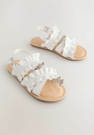 Baltas sandales ar viļņotām siksnām, mīkstu beigu krāsas zolīti un vienkāršu sprādzi aizdarei. Dzīvokļu zole ar vieglu teksturētu apdari.