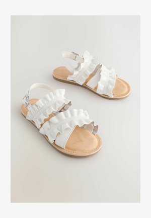 Valged sandaalid volditud rihmadega, pehme beež jalaosa ja lihtne pandla kinnitamine. Lame tall külgedel kergete tekstuuriliste detailidega.