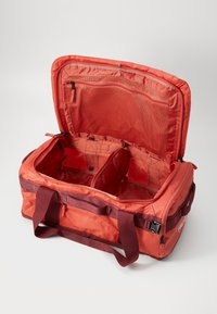 Red soft-sided duffel bag open with multiple compartments, mesh zipped pocket on lid, and maroon handles on white background.