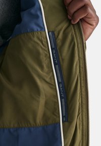 Interno della giacca GANT con tessuto verde oliva e blu navy, una trama liscia, una cerniera bianca visibile e dettagli con marchio sul rivestimento.