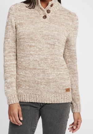 Žena v béžovém svetr z melírované pleteniny se třemi hnědými knoflíky u krku a tmavě šedých džínách střižených nad úroveň stehna.