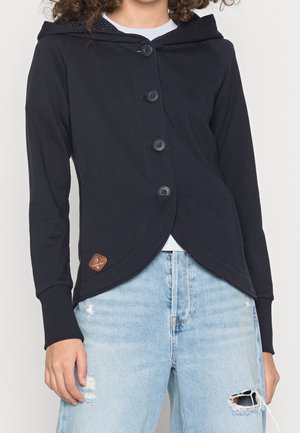 Mujer con sudadera con capucha negra abotonada y dobladillo curvado, y vaqueros desgastados azul claro con un desgarro en la rodilla derecha.