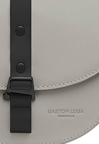 Ljusgrå läderväska med en böjd klaff, utrustad med ett svart rem och spänne, samt präglad "GASTON LUGA"-logotyp på ytan.